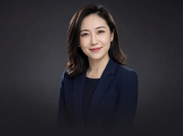 A professional headshot of a smiling woman in a navy blazer on a dark grey backdrop